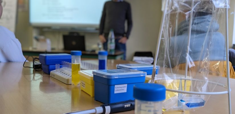 Das Bild zeigt ein Chemielabor. Im Hintergrund präsentiert ein Schüler seine Unterrichtsergebnisse.