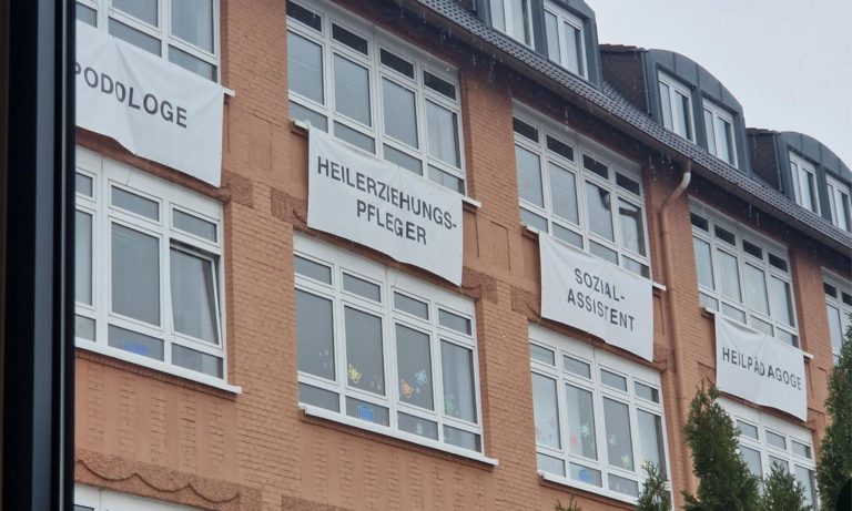 Man sieht ein Schulgebäude von außen. Klassische rote Backsteine und unter den Fenstern hängen Werbeplakate mit den unterschiedlichen Ausbildungsberufen der Schule.