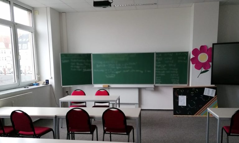 Man sieht einen leeren Klassenraum mit beschrifteter Tafel im Hintergrund.